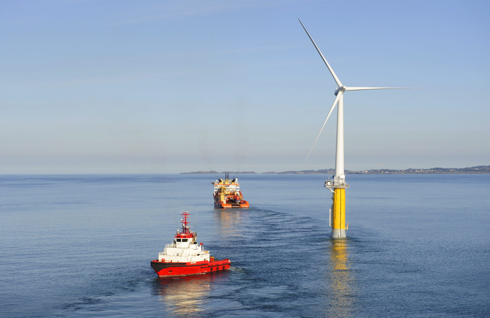 picture of an offshore wind turbine with two construction boats leaving from it, in separate directions.
